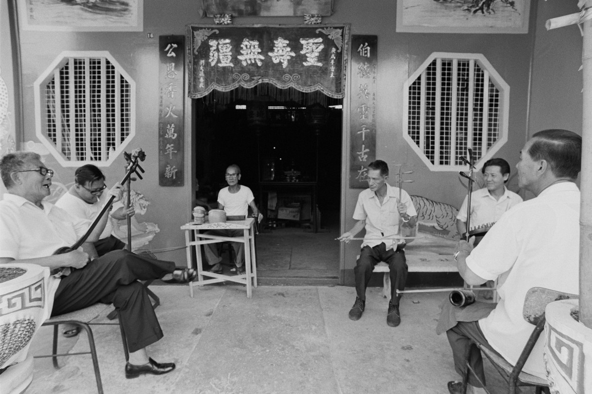 A group of seated senior men plays traditional Chinese instruments on a porch, overlooking ornate cultural decor, exuding a sense of communal gathering and cultural practice.