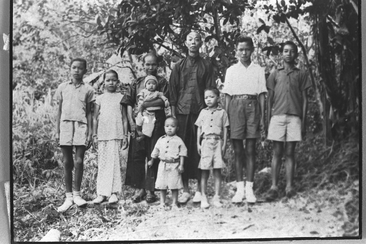 A group of eight individuals, possibly a family, stand together outdoors, with dense foliage forming the background. Most are looking directly at the camera.